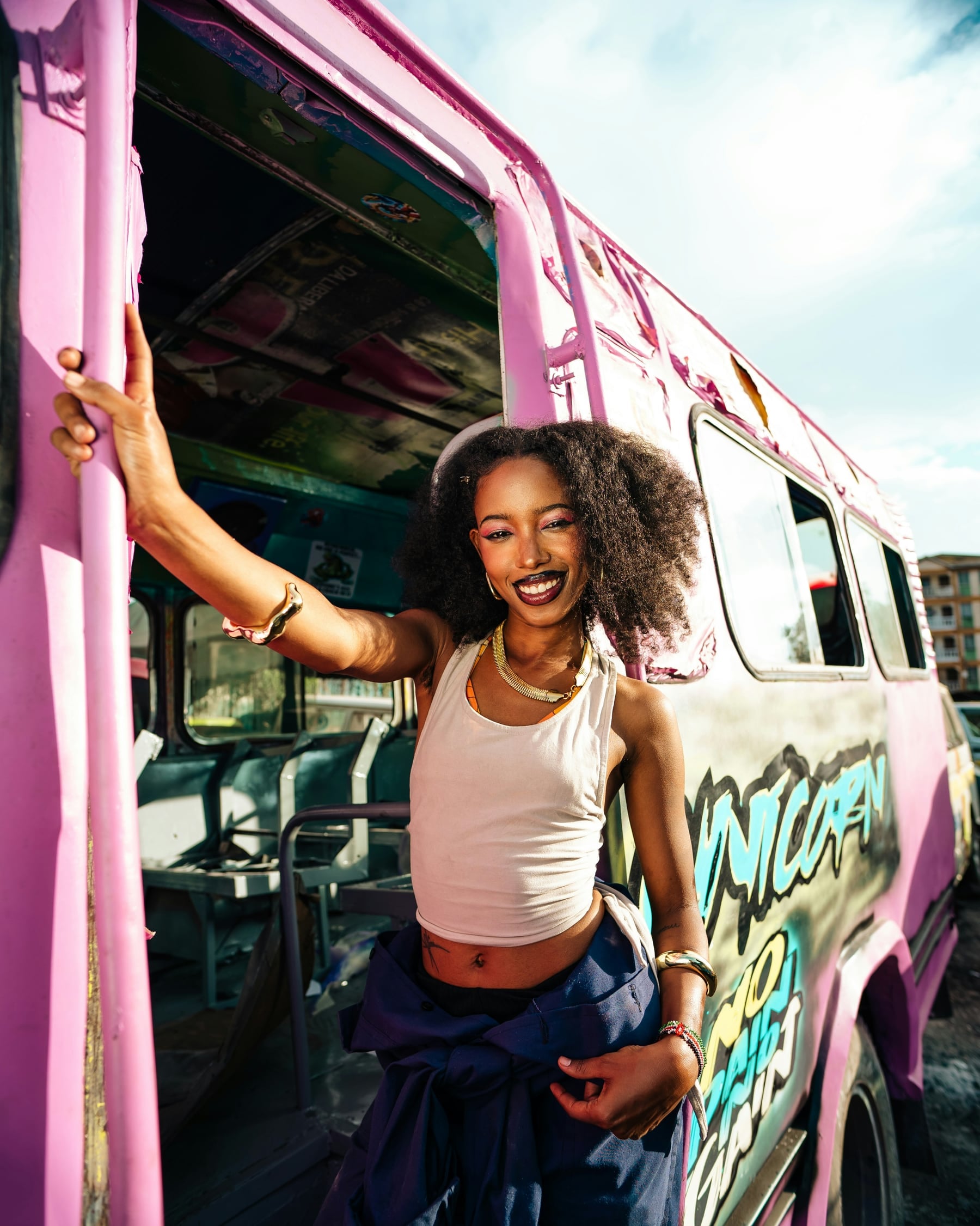 The woman from the first photograph stands in the doorway of the pink minibus in bright sun, smiling.