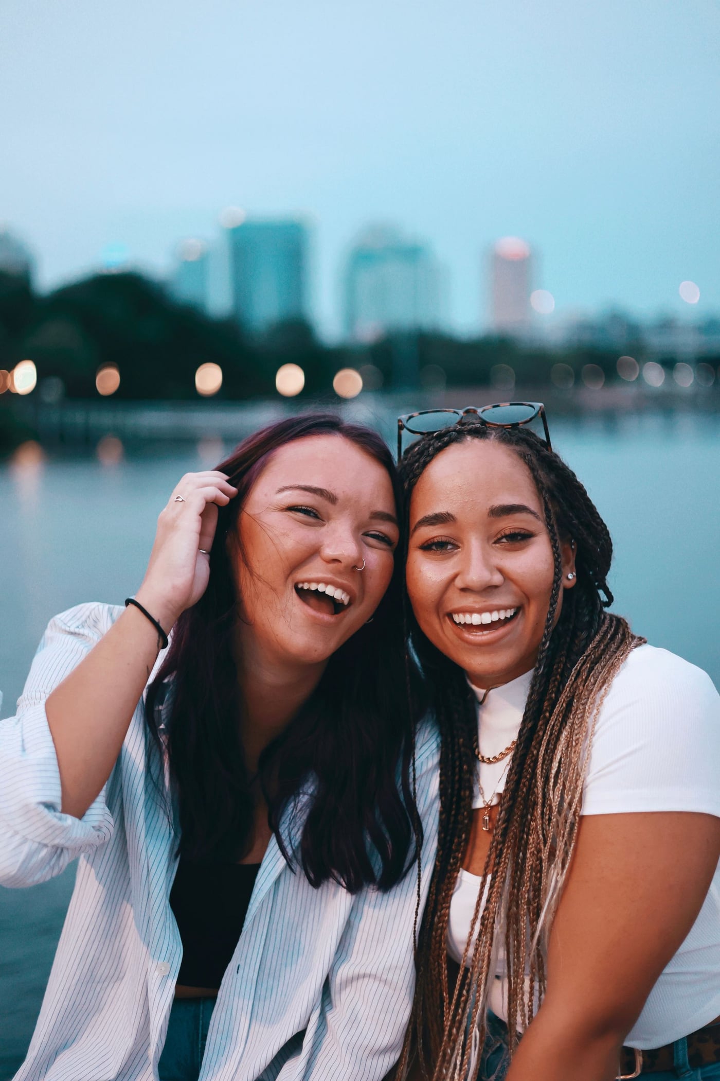 Two friends laughing together by the water at dusk, city lights behind them.