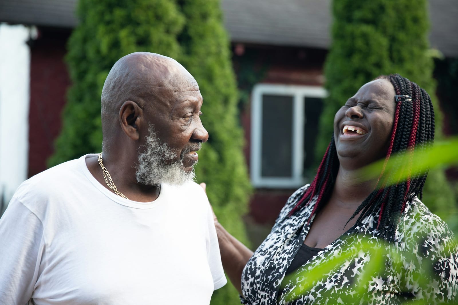 An older man with a white beard shares a laugh with his grown daughter outside a house.