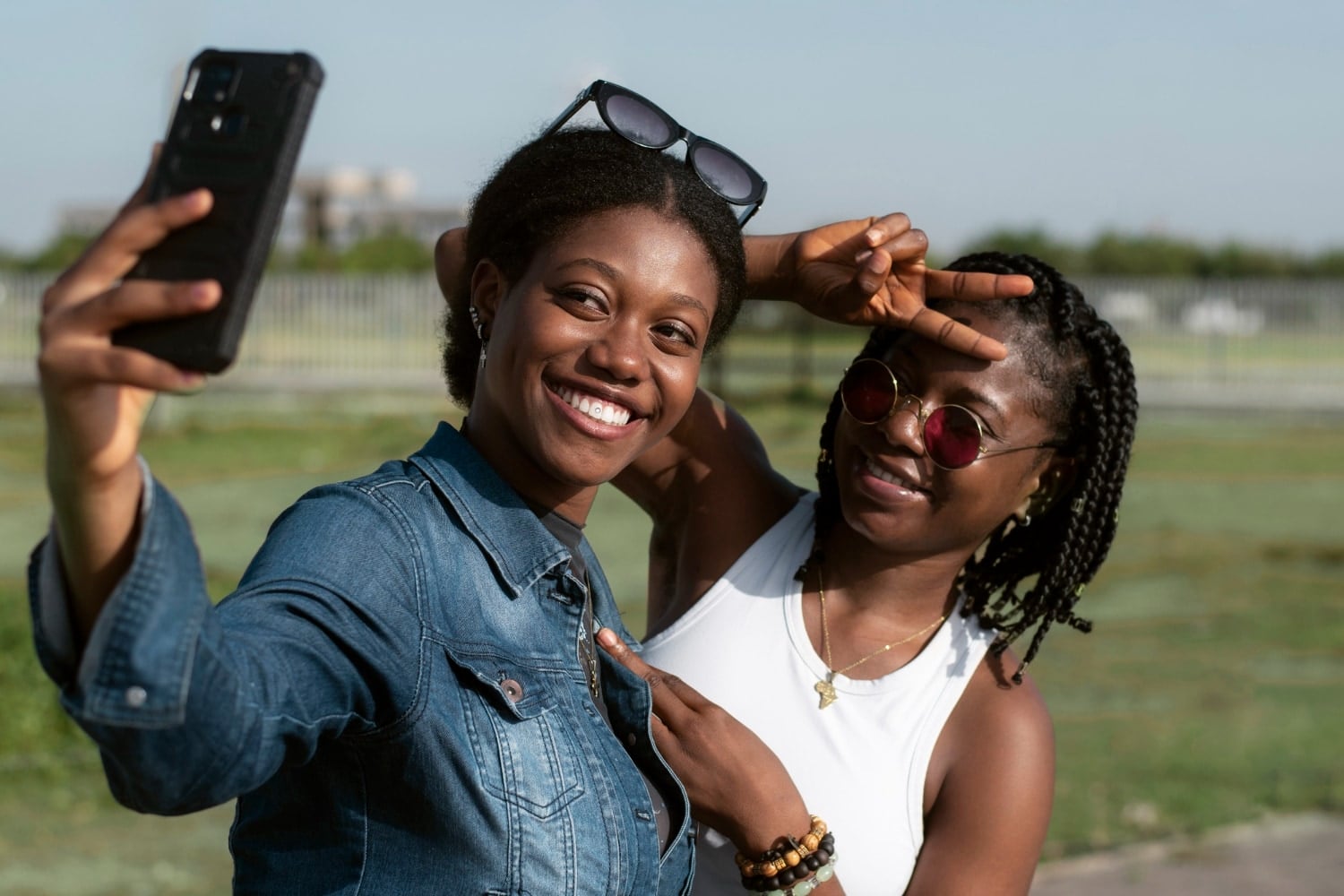 Two friends laughing as they take a selfie together in a sunny field.